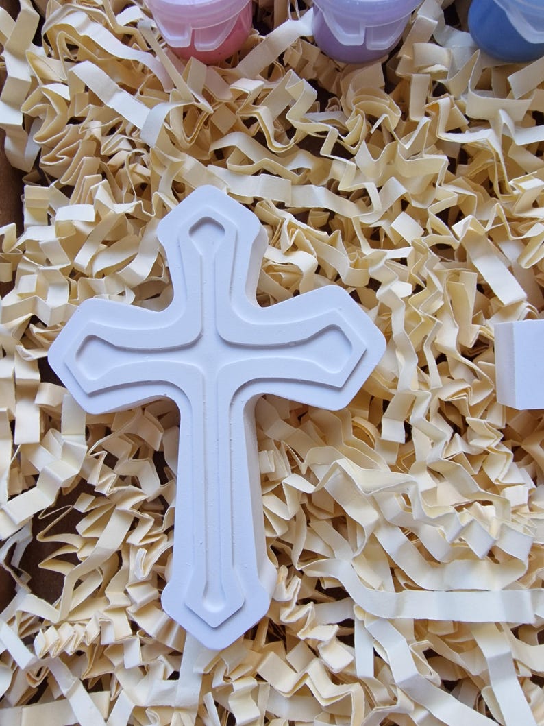 May include: White ceramic cross with a raised design. The cross is sitting on a bed of shredded paper.
