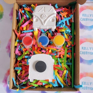 May include: A paint-your-own VW camper van craft kit. The kit includes two white ceramic camper vans, a paintbrush, and four small pots of paint: red, blue, yellow, and black.