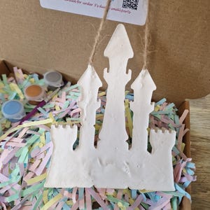 May include: A white castle-shaped craft kit with a string for hanging, inside a cardboard box filled with colorful paper shreds. Small paint pots and a brush are visible. The kit includes instructions and a QR code.
