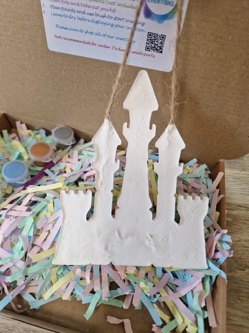 May include: A white castle-shaped craft kit with a string for hanging, inside a cardboard box filled with colorful paper shreds. Small paint pots and a brush are visible. The kit includes instructions and a QR code.