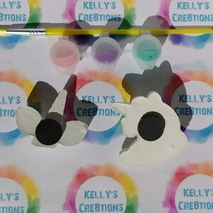 Puede incluir: Un kit de manualidades con dos imanes blancos con forma de unicornio, botes de pintura rosa, morada y turquesa, y un pincel con mango amarillo. El fondo incluye el texto "KELLY'S CREATIONS" en un c&iacute;rculo arco&iacute;ris.