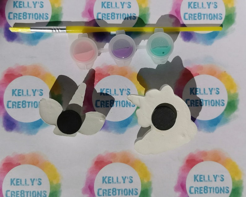 Puede incluir: Un kit de manualidades con dos imanes blancos con forma de unicornio, botes de pintura rosa, morada y turquesa, y un pincel con mango amarillo. El fondo incluye el texto "KELLY'S CREATIONS" en un c&iacute;rculo arco&iacute;ris.