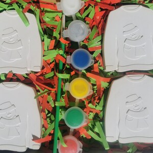 May include: Four white ceramic ornaments shaped like sweaters with a snowman design. The ornaments are surrounded by green and red shredded paper. There are five small paint pots with white, blue, yellow, green, and red paint.
