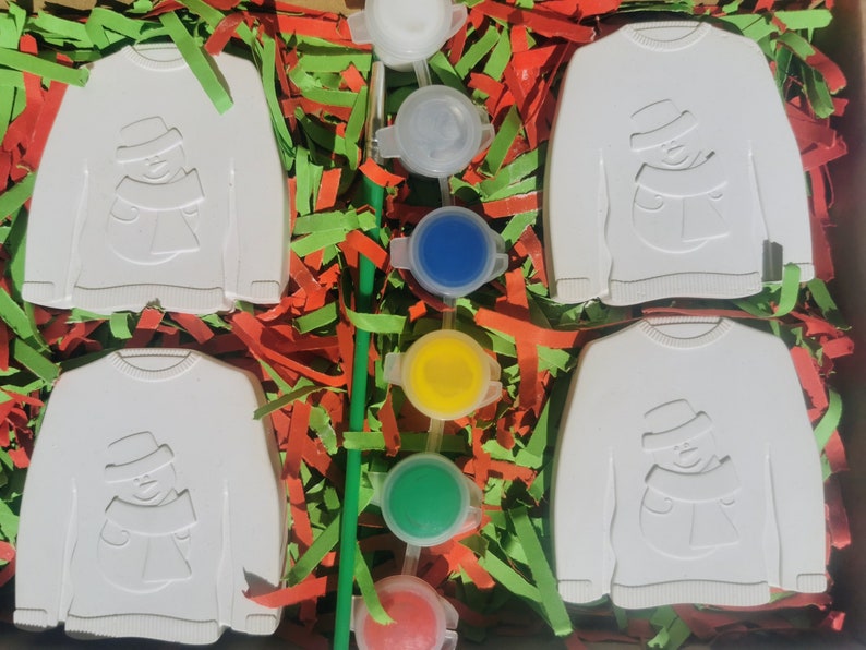 May include: Four white ceramic ornaments shaped like sweaters with a snowman design. The ornaments are surrounded by green and red shredded paper. There are five small paint pots with white, blue, yellow, green, and red paint.