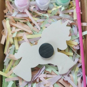 May include: A craft kit featuring a white unicorn-shaped magnet, three small paint pots in pink, purple, and green, and a pink paintbrush. The kit is nestled in colorful shredded paper inside a cardboard box.