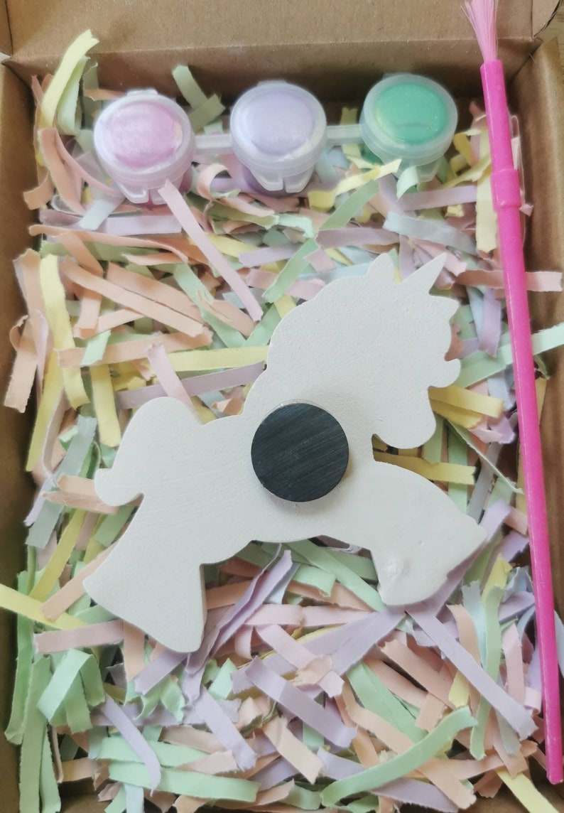 May include: A craft kit featuring a white unicorn-shaped magnet, three small paint pots in pink, purple, and green, and a pink paintbrush. The kit is nestled in colorful shredded paper inside a cardboard box.