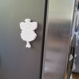May include: White 3D printed figurine of a fairy with a crown, wings, and a wand. The fairy is attached to a refrigerator with a magnet.