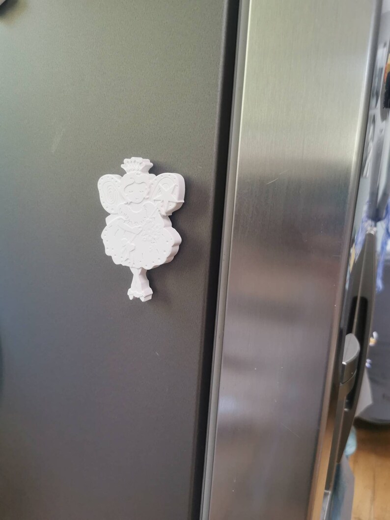 May include: White 3D printed figurine of a fairy with a crown, wings, and a wand. The fairy is attached to a refrigerator with a magnet.