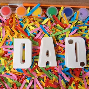 Op de afbeelding: Een knutselset met witte houten letters die "DAD" spellen en zes kleine verfpotjes met rode, oranje, gele, groene, blauwe en paarse verf. De letters en verfpotjes zijn omringd door kleurrijk versnipperd papier.