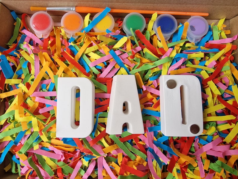 Op de afbeelding: Een knutselset met witte houten letters die "DAD" spellen en zes kleine verfpotjes met rode, oranje, gele, groene, blauwe en paarse verf. De letters en verfpotjes zijn omringd door kleurrijk versnipperd papier.