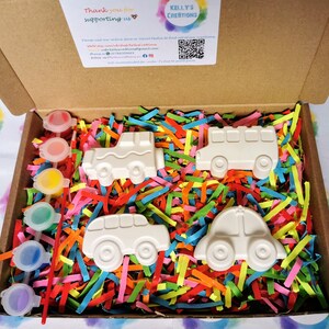 May include: A cardboard box filled with colorful shredded paper and four white ceramic vehicles. The vehicles are a bus, a car, a van, and a jeep. The box also contains six small bottles of paint.