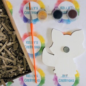 May include: A white wooden fox cutout with a magnet on the back. The fox is ready to be painted with the included paint pots: yellow, black, and brown. The paint pots are labeled "Kelly's Creations".