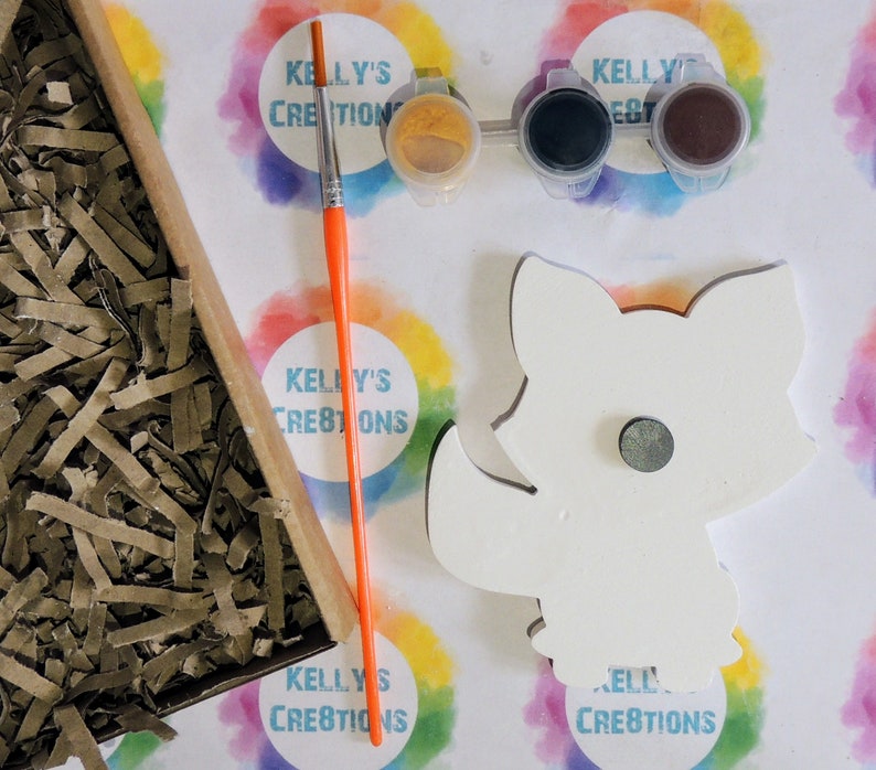 May include: A white wooden fox cutout with a magnet on the back. The fox is ready to be painted with the included paint pots: yellow, black, and brown. The paint pots are labeled "Kelly's Creations".