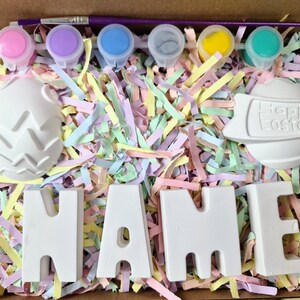 May include: A craft kit for painting ceramic Easter decorations. The kit includes six small paint pots in pink, purple, blue, gray, yellow, and green, a paintbrush, and five ceramic pieces: two eggs, one with a textured surface, one with the words "Happy Easter", and three letters spelling "NAME".