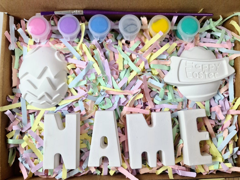 May include: A craft kit for painting ceramic Easter decorations. The kit includes six small paint pots in pink, purple, blue, gray, yellow, and green, a paintbrush, and five ceramic pieces: two eggs, one with a textured surface, one with the words "Happy Easter", and three letters spelling "NAME".
