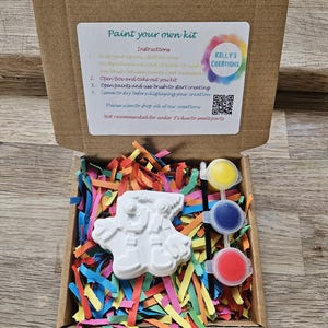 May include: A paint-your-own kit in a cardboard box, containing a white ceramic figure, three paint pots (yellow, blue, and red), and colourful paper shreds. The box includes instructions and the text "KELLY'S CREATIONS".