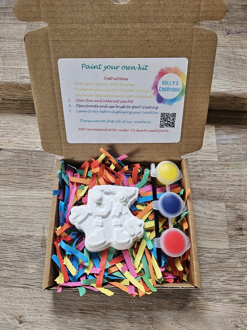 May include: A paint-your-own kit in a cardboard box, containing a white ceramic figure, three paint pots (yellow, blue, and red), and colourful paper shreds. The box includes instructions and the text "KELLY'S CREATIONS".