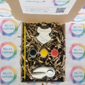May include: A craft kit with a Ravenclaw crest, paint pots in black, yellow, and red, a paintbrush, and a ceramic item. The kit is in a cardboard box with shredded paper filler. The box is surrounded by stickers with the text "Kelly's Creations".