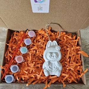 May include: A craft kit in a cardboard box, featuring a white ceramic bunny figure with a pumpkin face, surrounded by orange paper shreds. The kit includes small paint pots in various colors and a paintbrush. The box has a label that says "Kelly's Creations".