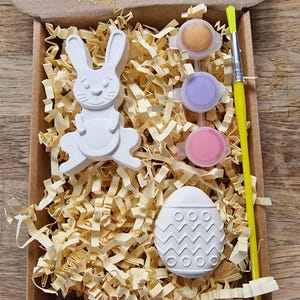 May include: A craft kit for painting a ceramic bunny and egg. The kit includes three small pots of paint in orange, purple, and pink, a paintbrush, and a small piece of shredded paper.