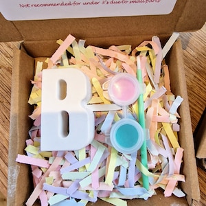 May include: A white ceramic letter B with two small paint pots, one pink and one turquoise, and a paintbrush. The letter and paint pots are sitting on a bed of colourful shredded paper in a cardboard box. The box has a label that says "Open paints and use brush to start creating. Leave to dry before displaying your creation. Please scan to shop all of our creations. Not recommended for under 3's due to small parts."