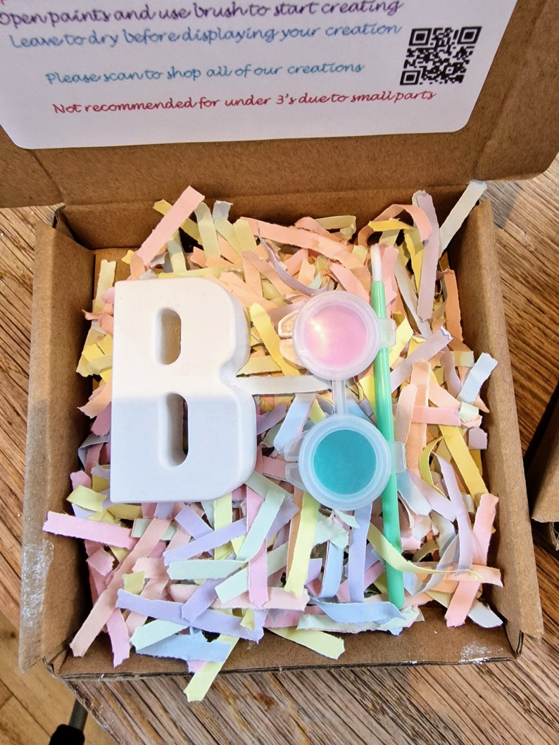 May include: A white ceramic letter B with two small paint pots, one pink and one turquoise, and a paintbrush. The letter and paint pots are sitting on a bed of colourful shredded paper in a cardboard box. The box has a label that says "Open paints and use brush to start creating. Leave to dry before displaying your creation. Please scan to shop all of our creations. Not recommended for under 3's due to small parts."