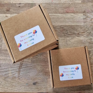 May include: Two small, brown cardboard boxes with white labels. The labels read "Thank you for coming to my party" with the names "Ethan" and "Liam" below. Colorful balloon graphics are also on the labels.