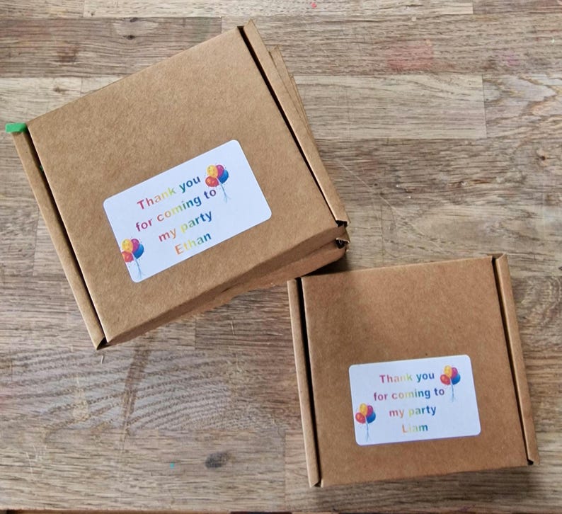 May include: Two small, brown cardboard boxes with white labels. The labels read "Thank you for coming to my party" with the names "Ethan" and "Liam" below. Colorful balloon graphics are also on the labels.