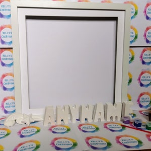 May include: White wooden frame with a white blank canvas inside.  Below the frame are white letters spelling out "ANYNAME" and a rainbow and star made of white plaster.  There are also small bottles of paint and a paintbrush on a white surface with a repeating pattern of colorful circles.