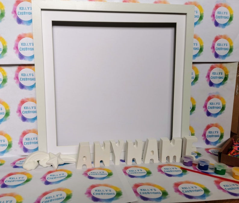 May include: White wooden frame with a white blank canvas inside.  Below the frame are white letters spelling out "ANYNAME" and a rainbow and star made of white plaster.  There are also small bottles of paint and a paintbrush on a white surface with a repeating pattern of colorful circles.