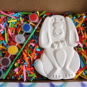 May include: A white ceramic dog figurine with a bow tie, paint pots in red, blue, yellow, brown, and black, and a green paintbrush in a cardboard box filled with colorful shredded paper.