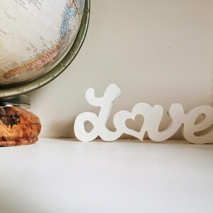 May include: White wooden sign spelling out the word "love" with a heart in the "o". The sign is sitting on a white surface.