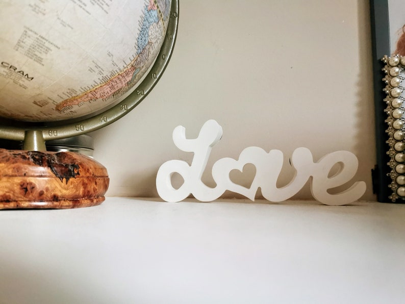 May include: White wooden sign spelling out the word "love" with a heart in the "o". The sign is sitting on a white surface.