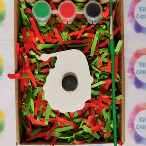 May include: A white ceramic Santa Claus ornament with a hole in the center, surrounded by red and green shredded paper. Three small paint pots with green, red, and black paint are included, along with a paintbrush. The box is labeled "Kelly's Creations".