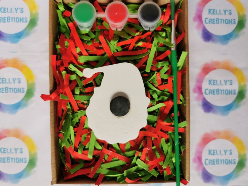 May include: A white ceramic Santa Claus ornament with a hole in the center, surrounded by red and green shredded paper. Three small paint pots with green, red, and black paint are included, along with a paintbrush. The box is labeled "Kelly's Creations".