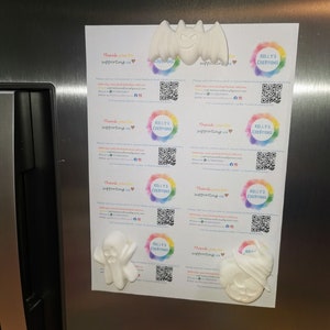 May include: Four white ceramic Halloween-themed ornaments, including a bat, ghost, pumpkin, and a ghost with a hat. The ornaments are attached to a refrigerator with magnets. The ornaments are blank and ready to be painted. The background is a stainless steel refrigerator.