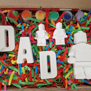 May include: A craft kit in a cardboard box, featuring the word "DAD" in white block letters, white toy figures, and small paint pots with a paintbrush. The box is filled with colorful shredded paper.