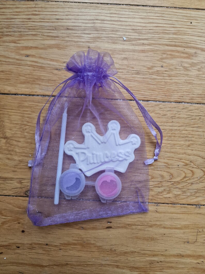 May include: A purple mesh bag containing a white crown-shaped craft with the word "Princess" on it, two small containers of paint, and a paintbrush.