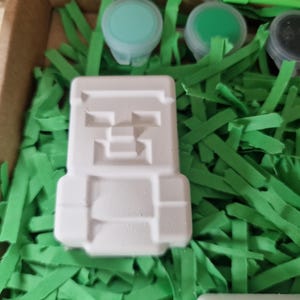 Puede incluir: Una figura blanca y cuadrada con un dise&ntilde;o pixelado, posiblemente un personaje de videojuego, se encuentra entre hierba de papel verde. Se ven peque&ntilde;os botes de pintura en tonos verdes y azules, lo que sugiere una actividad de manualidades o pintura.