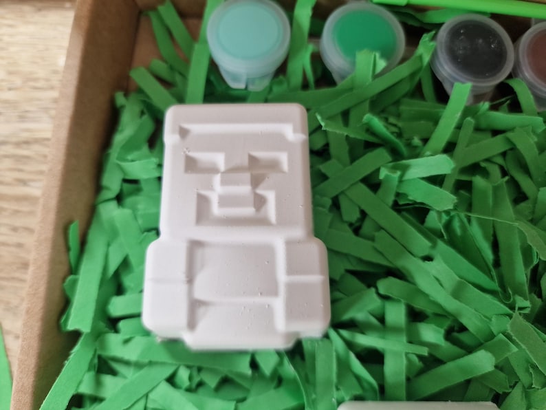 Puede incluir: Una figura blanca y cuadrada con un dise&ntilde;o pixelado, posiblemente un personaje de videojuego, se encuentra entre hierba de papel verde. Se ven peque&ntilde;os botes de pintura en tonos verdes y azules, lo que sugiere una actividad de manualidades o pintura.
