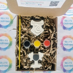May include: A craft kit in a cardboard box, featuring a white Slytherin crest and a trophy. The kit includes three paint pots in black, yellow, and red, along with a yellow paintbrush. The box is filled with brown paper shreds.