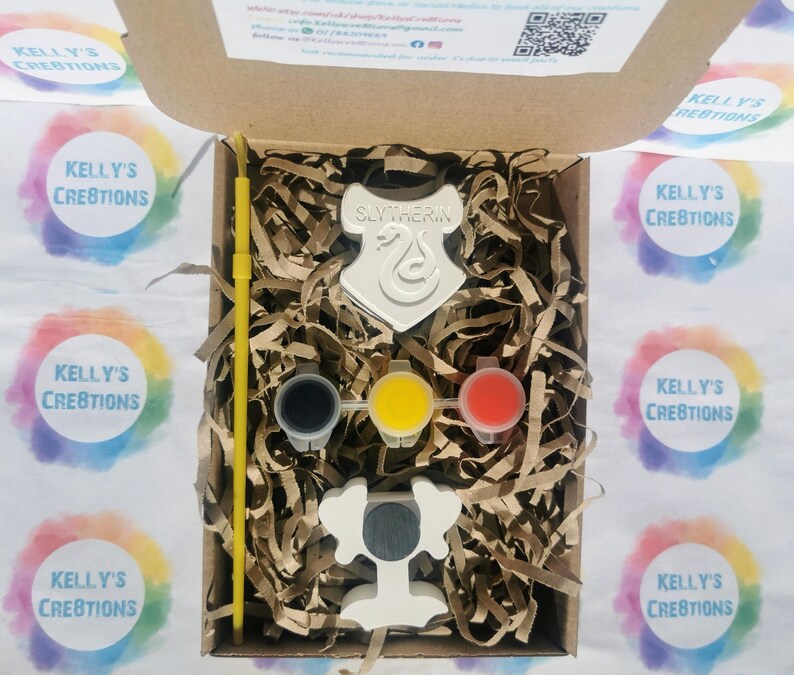 May include: A craft kit in a cardboard box, featuring a white Slytherin crest and a trophy. The kit includes three paint pots in black, yellow, and red, along with a yellow paintbrush. The box is filled with brown paper shreds.