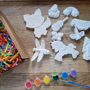 May include: A set of 10 white ceramic figurines, including a fox, a dragonfly, a unicorn, a butterfly, a parrot, a rainbow, a violin, a crown, and a princess. The figurines are in a box with colorful shredded paper. There are also 7 small paint pots with different colors.