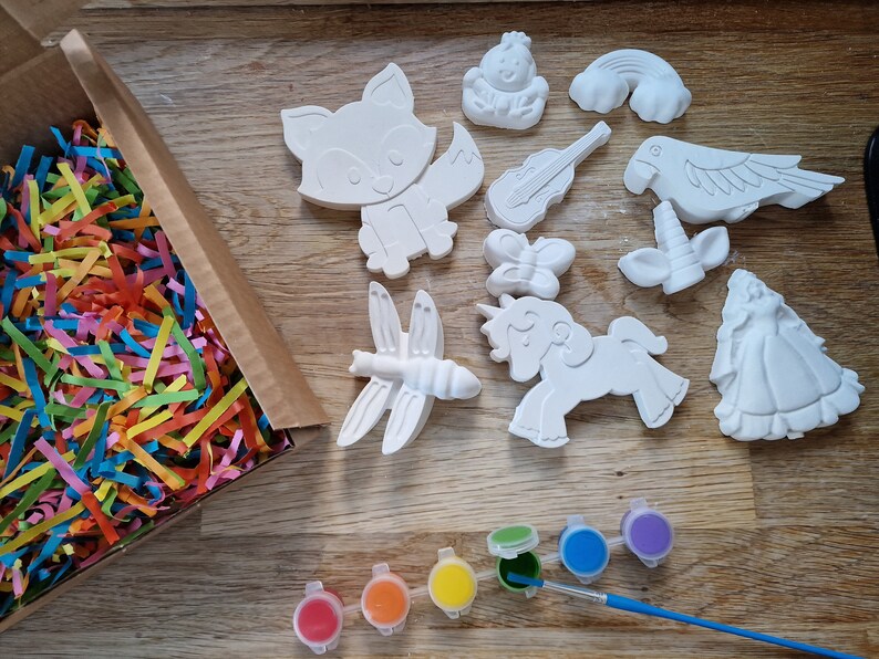 May include: A set of 10 white ceramic figurines, including a fox, a dragonfly, a unicorn, a butterfly, a parrot, a rainbow, a violin, a crown, and a princess. The figurines are in a box with colorful shredded paper. There are also 7 small paint pots with different colors.