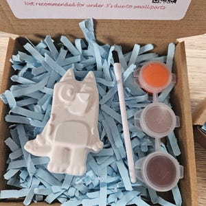 May include: A craft kit in a cardboard box, containing a white plaster dog figurine, paint pots, and a paintbrush. The box is filled with blue paper shreds. Text on the box reads: "Open paints and use brush to start creating."