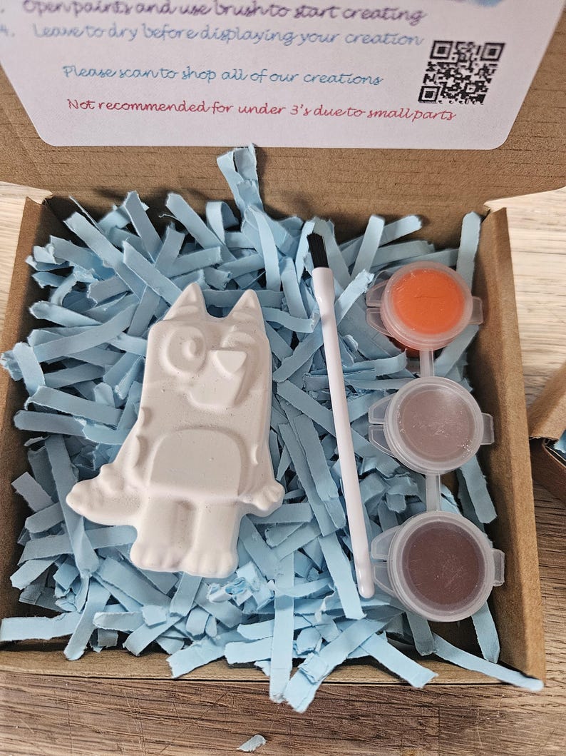May include: A craft kit in a cardboard box, containing a white plaster dog figurine, paint pots, and a paintbrush. The box is filled with blue paper shreds. Text on the box reads: "Open paints and use brush to start creating."