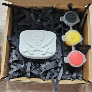 May include: A cardboard box filled with black paper shreds containing a white rectangular object embossed with a skull and crossbones design, accompanied by three small circular containers of black, yellow, and red paint and a thin black paintbrush.