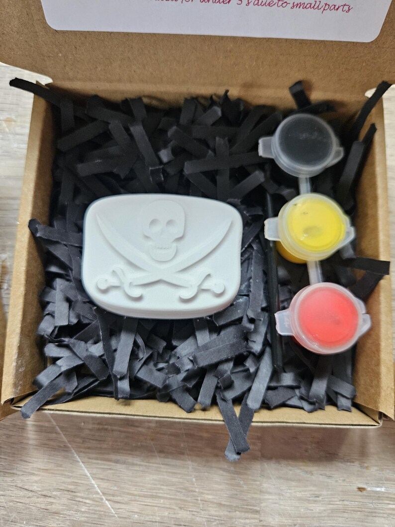 May include: A cardboard box filled with black paper shreds containing a white rectangular object embossed with a skull and crossbones design, accompanied by three small circular containers of black, yellow, and red paint and a thin black paintbrush.