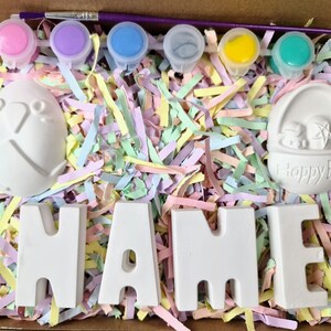 May include: A craft kit with white ceramic Easter-themed figurines, including a bunny, an egg, and the word "NAME". The kit also includes six small paint pots with different colors: pink, purple, blue, gray, yellow, and green.