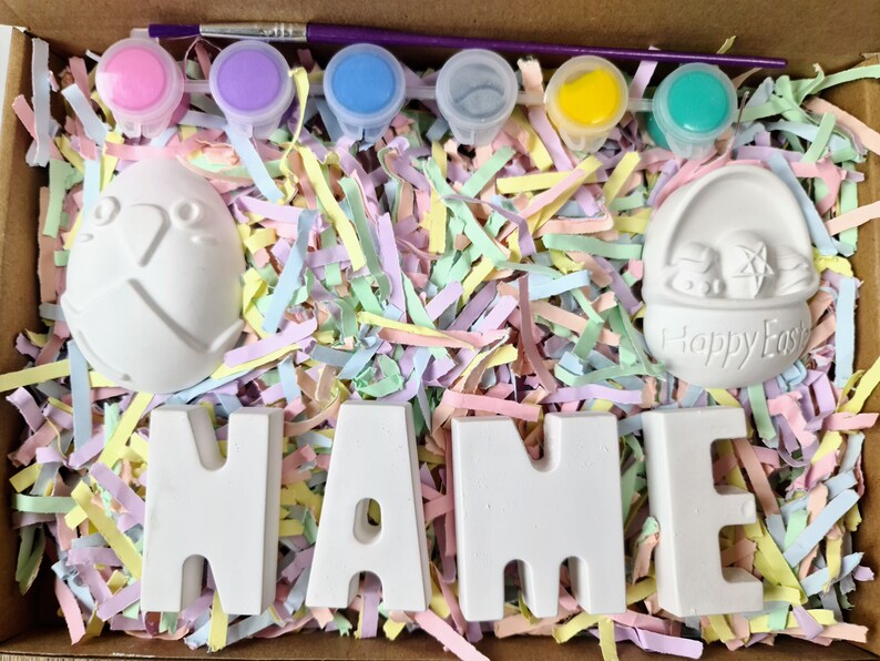 May include: A craft kit with white ceramic Easter-themed figurines, including a bunny, an egg, and the word "NAME". The kit also includes six small paint pots with different colors: pink, purple, blue, gray, yellow, and green.